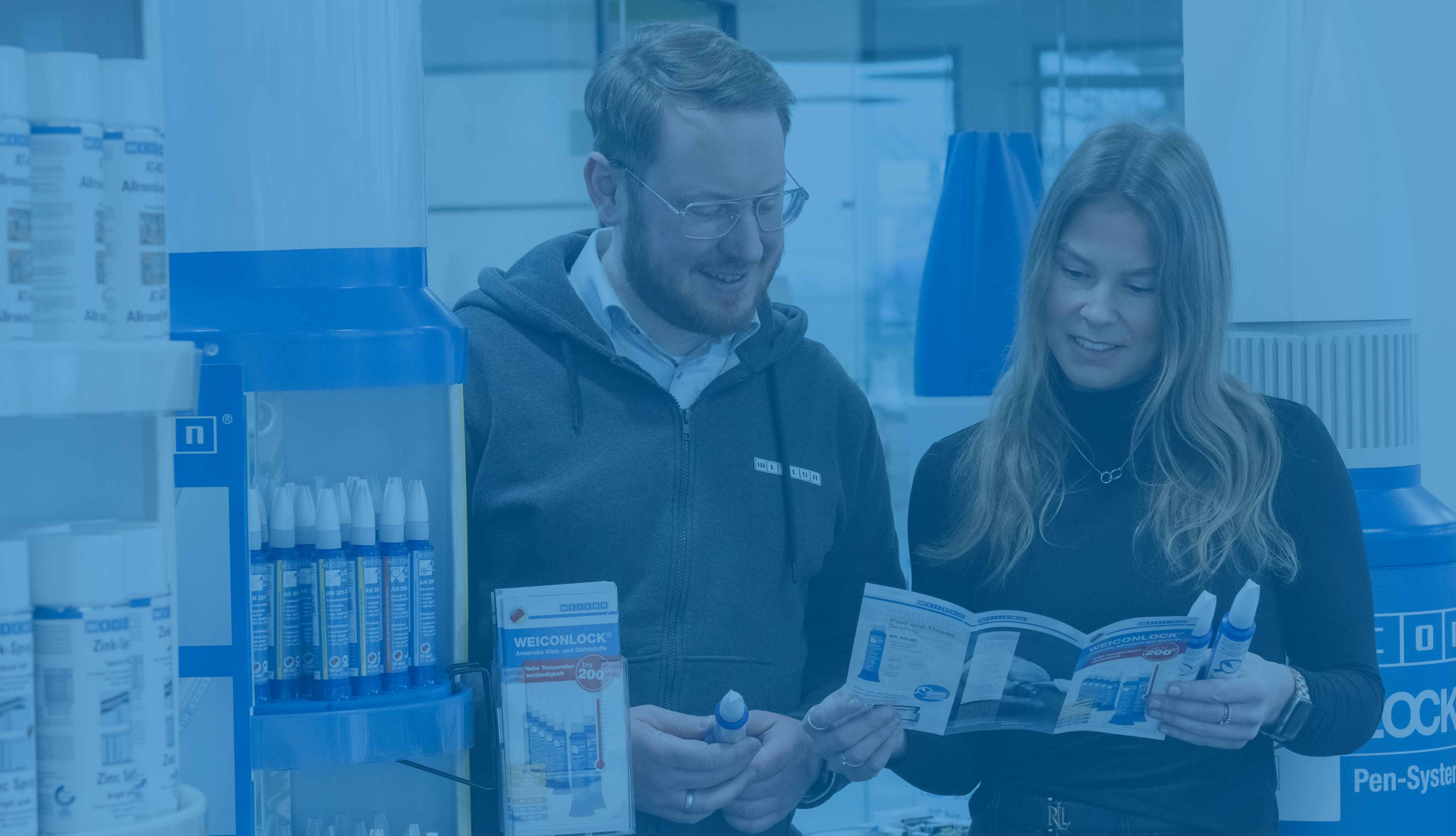 Ein Mann und eine Frau stehen nebeneinander in einem Raum und schauen sich einen Weicon Flyer an, neben ihnen stehen Verkaufsdisplay von Weicon. Das Bild ist blau hinterlegt