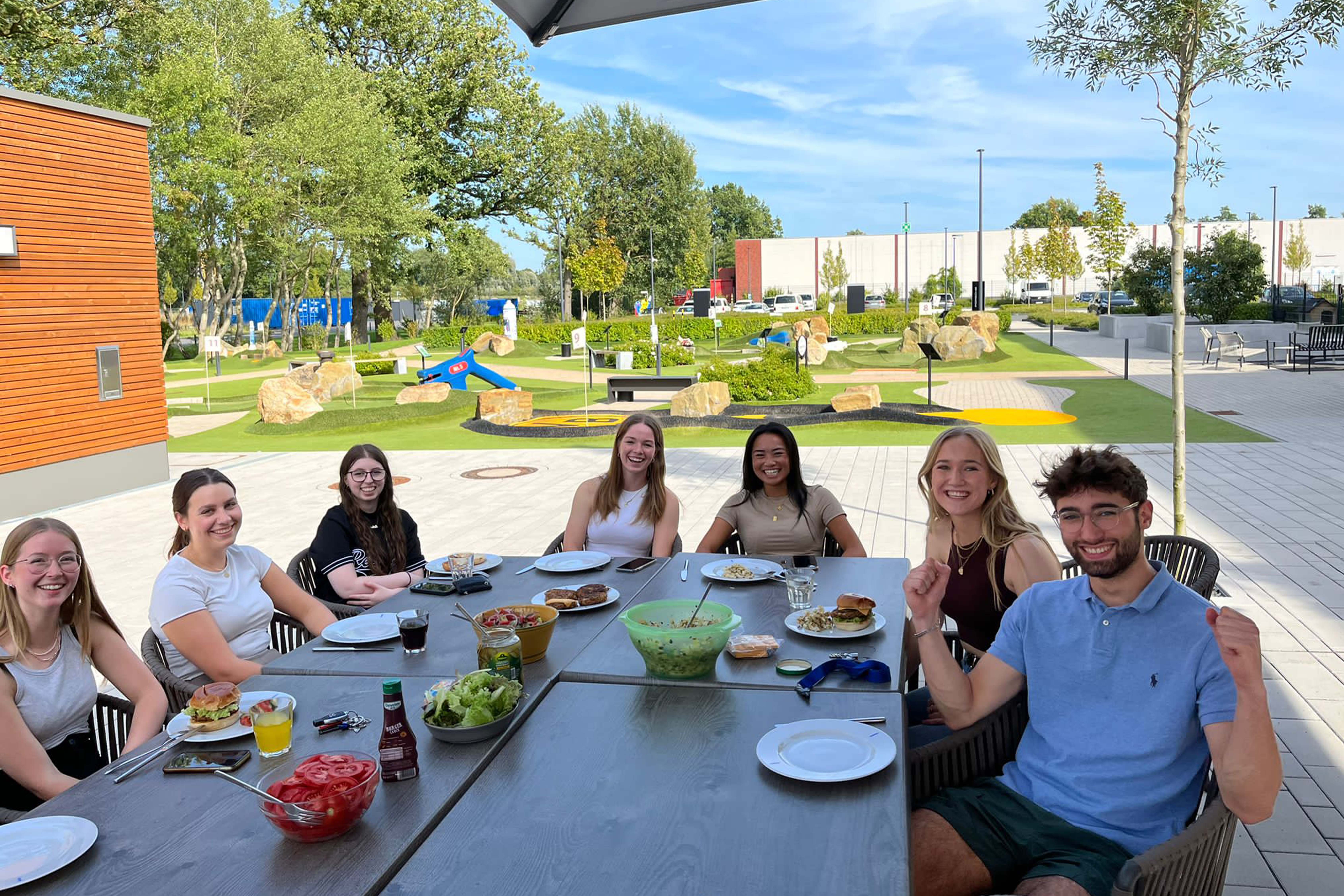 Gruppe von Jugendlichen sitzt an einem Tisch, alle gucken in die Kamera, Essen steht auf dem Tisch, im Hintergrund ist ein Minigolfplatz