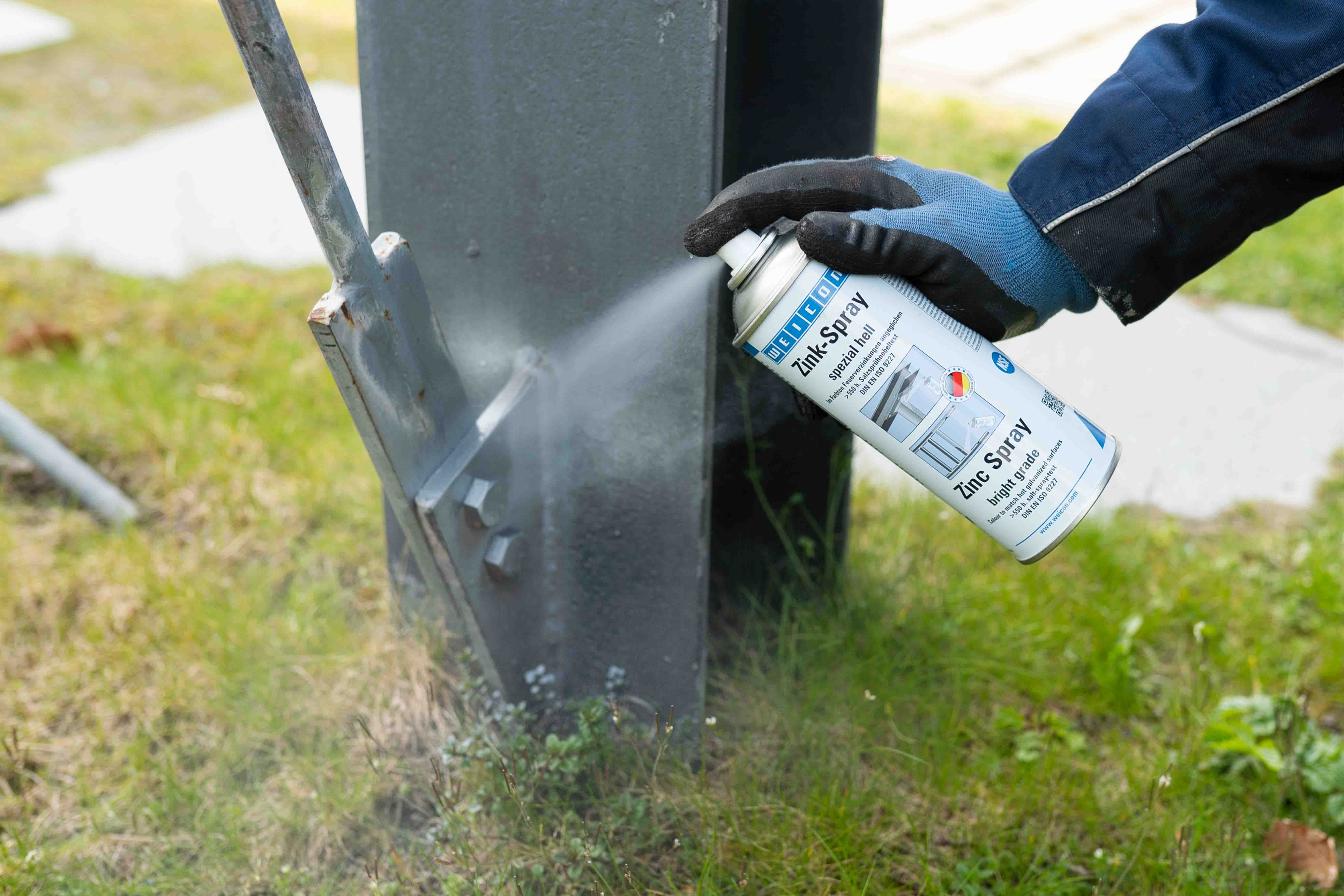 Metallpfahl im Gras wird unten an Schauben mit Weicon Zink-Spray hell besprüht