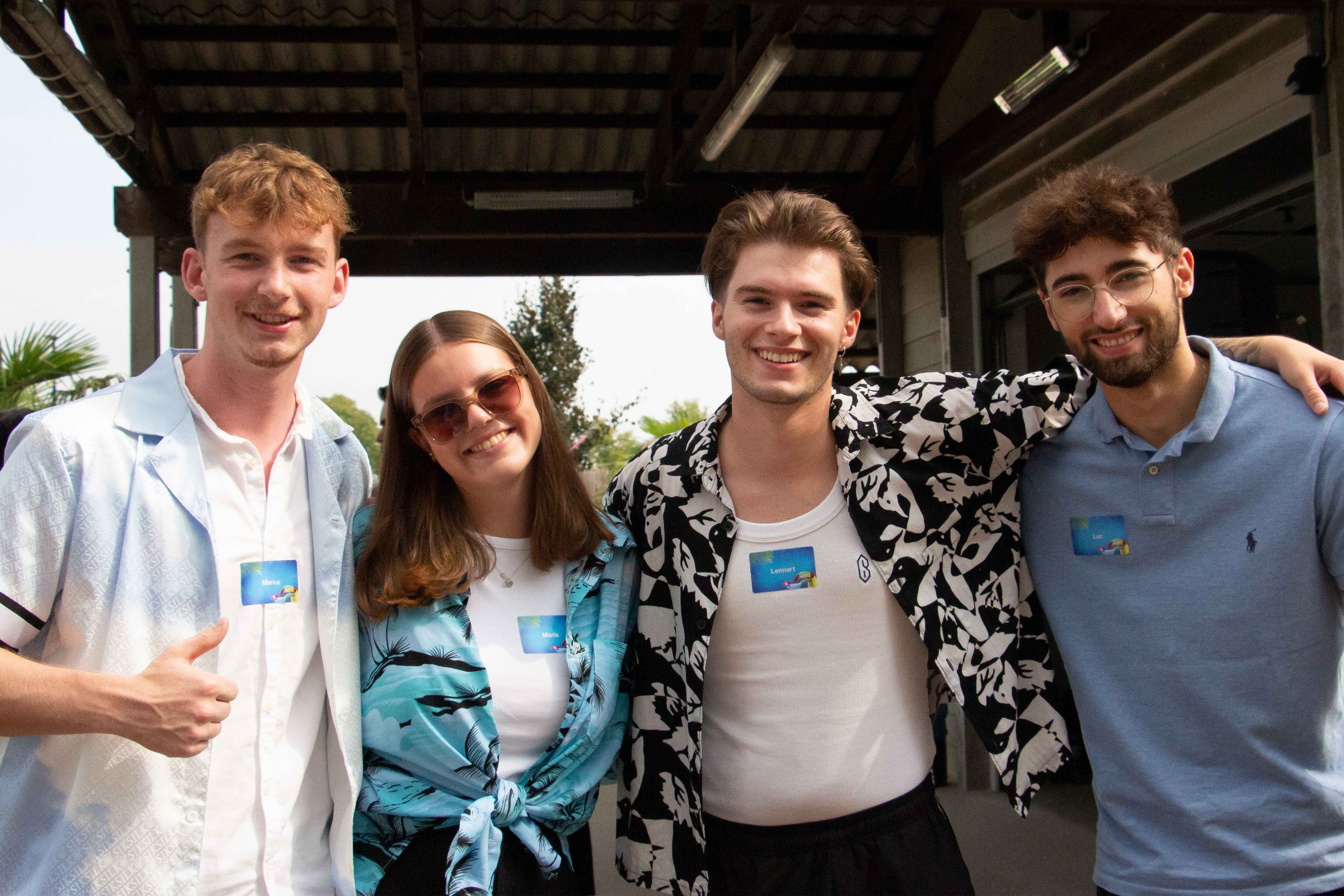 3 junge Männer und eine junge Frau, die in einer Reihe stehen, sich in den Arm nehmen und lachen