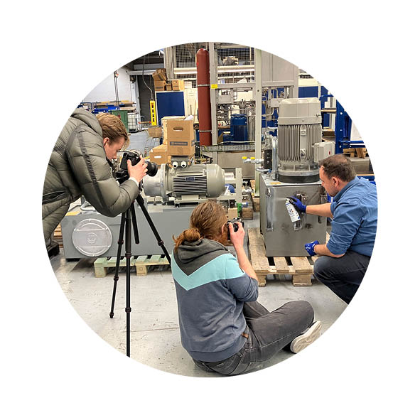 Drei Männer sind in einer Halle, steht an einem Stativ und fotografiert, einer sitzt auf dem Boden und fotografiert und der Dritte bückt sich und sprüht etwas auf eine Anlage