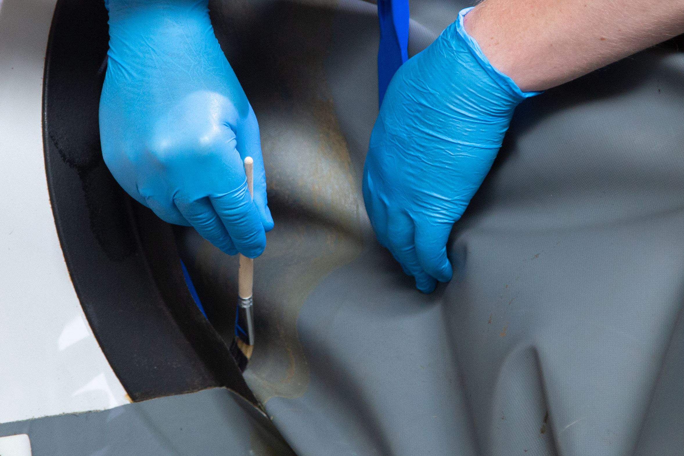 GMK 2510_Gummi-Metall-Kleber_Boot-reparieren Kleber wird auf ein Teil eines Gummibootes mit einem Pinsel und blauen Handschuhen gestrichen