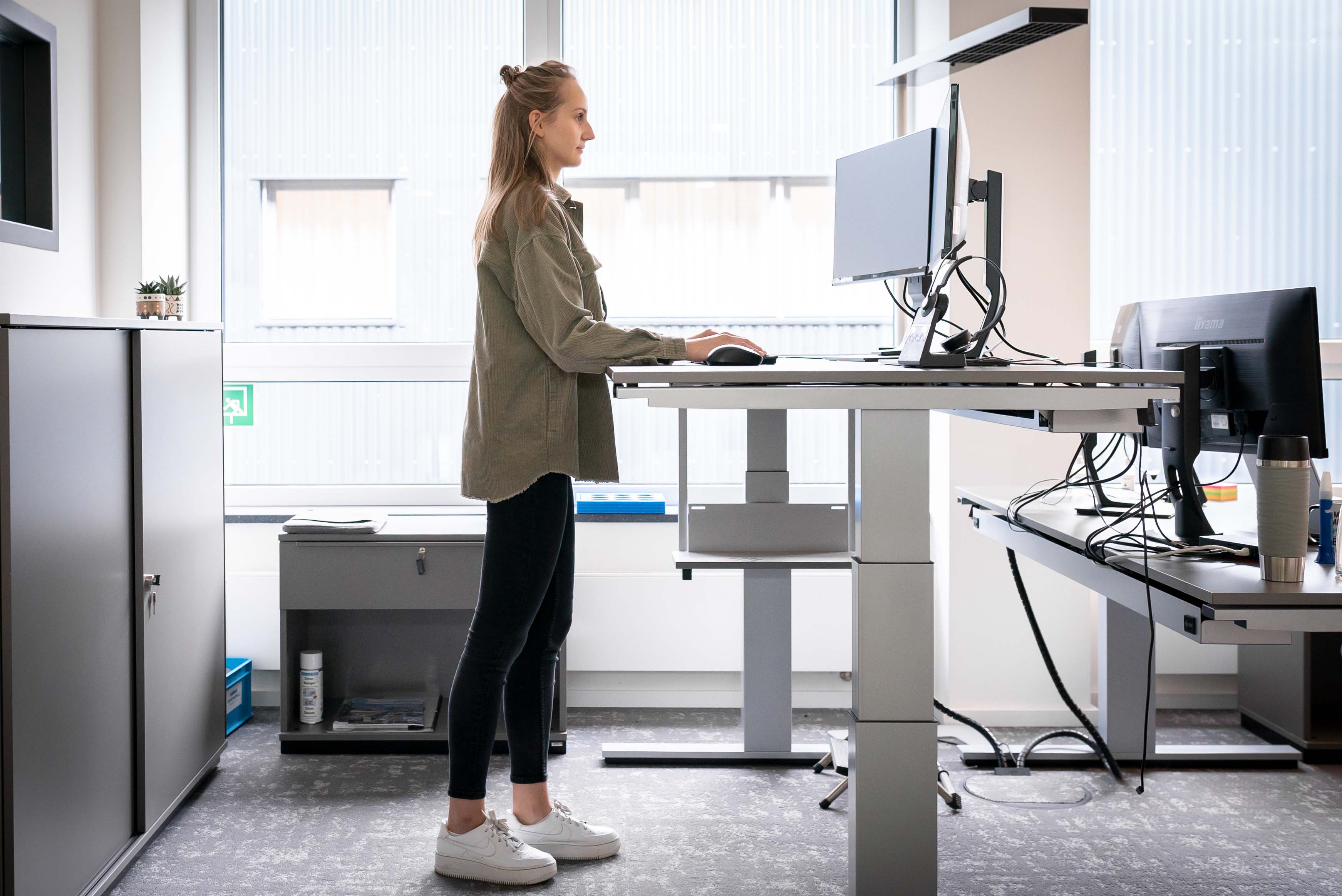 eine junge Frau steht in einem Büro an einem höhenverstellbaren Schreibtisch und arbeitet