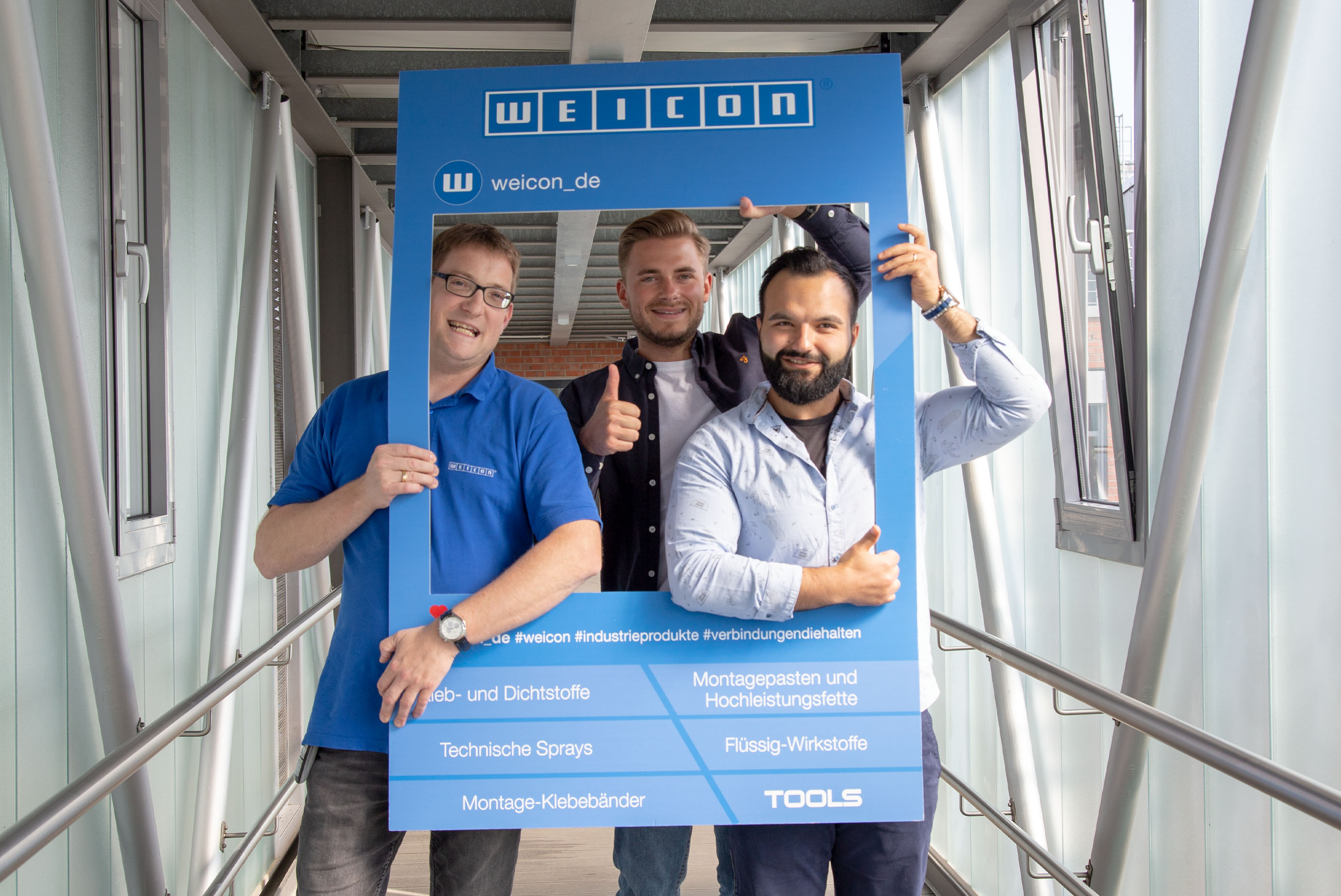 Drei Männer stehen in einem Weicon-Rahmen, halten ihn fest und lachen in die Kamera