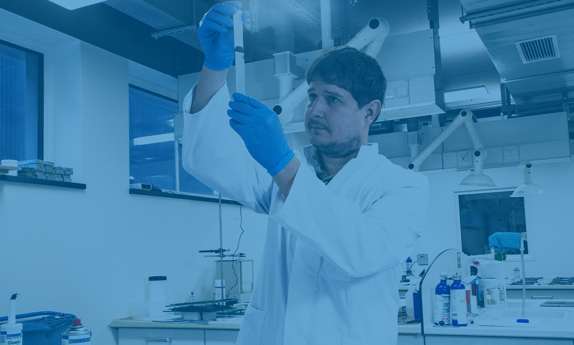 Ein Mann steht mit einem Kittel und blauen Handschuhen in einem Labor und hält ein Reagenzglas hoch, das Bild ist blau hinterlegt