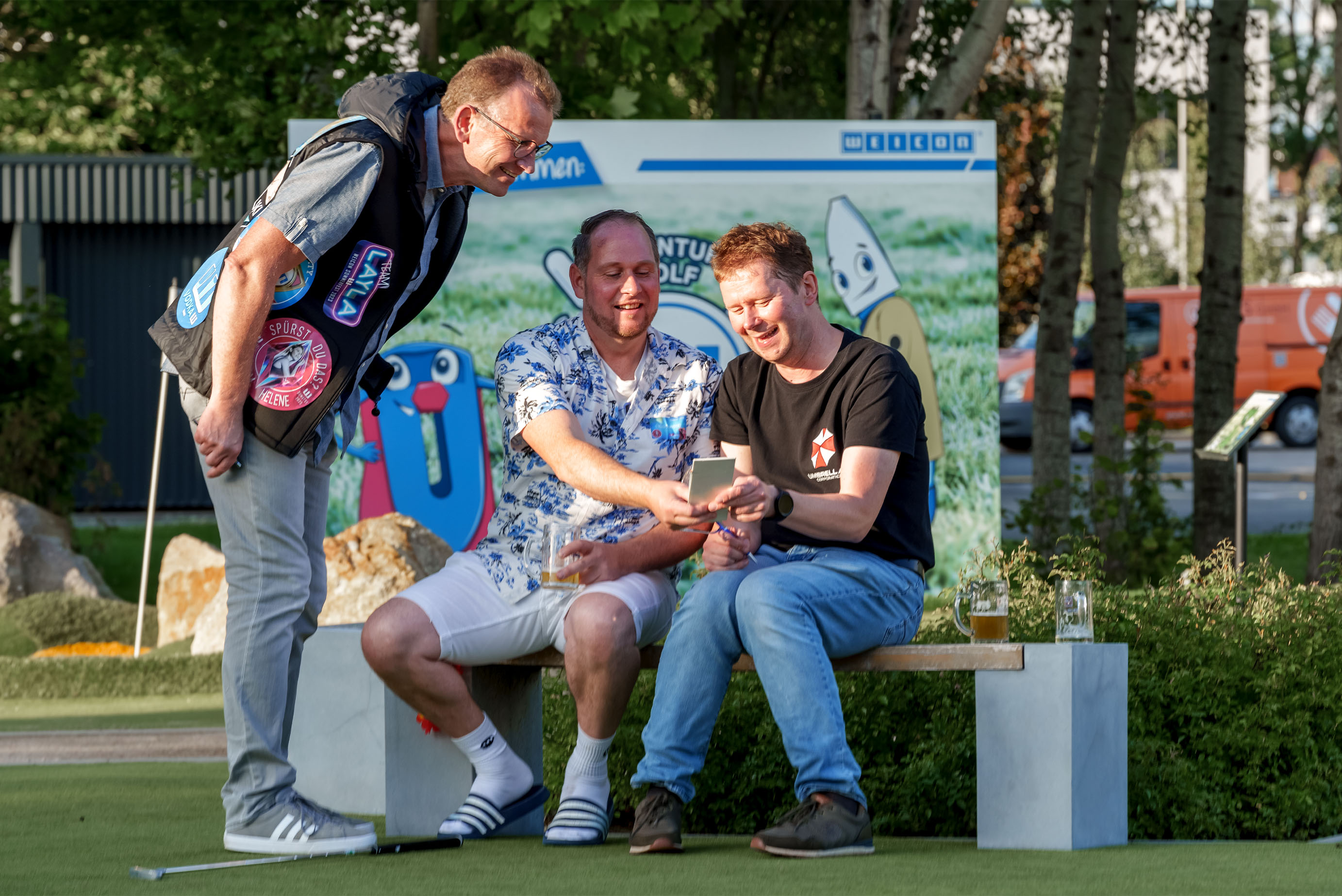 Zwei Männer sitzen auf einer Bank und schauen sich etwas auf dem Handy an, ein weiterer Mann steht daneben und guckt auch lackend auf das Handy